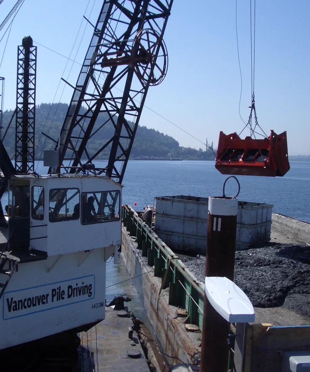 A crane on a dredge barge lifts a clamshell bucket over the water with a Vancouver Pile Driving vessel alongside